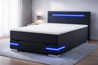 Zwart gestoffeerd boxspringbed met geïntegreerde blauwe LED-verlichting in hoofdbord en onderzijde, diagonaal geplaatst in een rustige, moderne slaapkamer met grijze tinten en zacht daglicht.